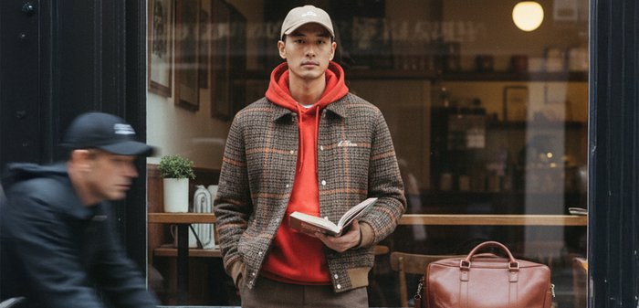 Barna kockás dzsekiben és piros kapucnis pulcsiban álló férfi könyvet olvas egy kávézó ablaka előtt, közelében egy barna bőr táska.