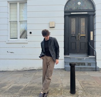 Giacca di pelle nera su una maglietta nera, pantaloni beige a pieghe, scarpe di pelle scure, in piedi vicino a una colonna nera accanto a un edificio bianco.