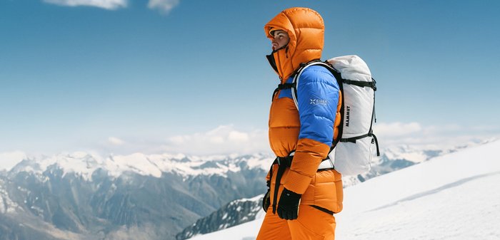 Oransje og blå puffjakke med teksturert stoff, brukt med svarte hansker og utstyrt med en hvit ryggsekk med stropper og logo.