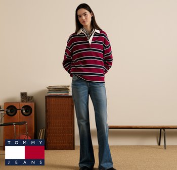 Femme debout les mains dans les poches, portant un polo rugby rayé bordeaux et un jean bleu, dans une pièce minimaliste avec des haut-parleurs vintage et un banc.