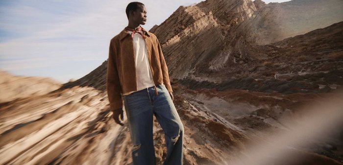 Junger Mann in brauner Jacke und zerrissener Jeans steht in felsigem, bergigem Gelände unter einem teilweise bewölkten Himmel.
