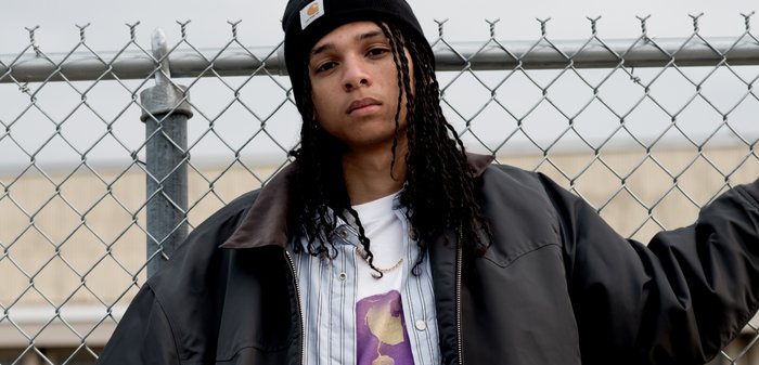 Joven con cabello largo trenzado que lleva un gorro negro y una chaqueta, de pie junto a una valla de cadena al aire libre.