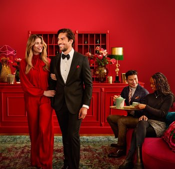 En mann i svart dress og en kvinne i rød bluse poserer sammen, mens to andre som sitter nyter te på et grønt og hvitt fat. Rød bakgrunn.