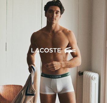 Boxer bianchi con un elastico verde che presenta il logo "LACOSTE", indossati da un uomo che tiene un indumento di colore chiaro, in un contesto minimalista.
