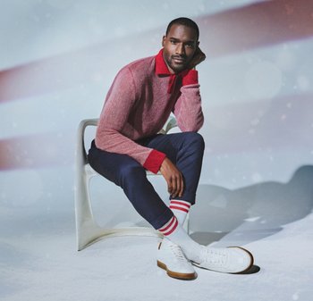 Roter und rosa gestrickter Pullover, marineblaue Hose, weiße Schuhe und gestreifte Socken. Sitzend auf einem weißen Stuhl vor einem weichen Hintergrund mit Bokeh-Effekten.