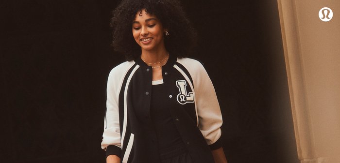 Giacca bomber bianca e nera con design varsity, con una patch a forma di lettera sul petto, indossata sopra una maglietta nera. Capelli ricci visibili.