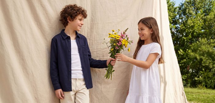 Garçon en chemise bleu marine et pantalon beige remet un bouquet de fleurs sauvages multicolores à une fille souriante en robe blanche sur fond beige.