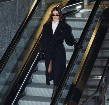 Czarny płaszcz z paskiem, z bocznym rozcięciem, w połączeniu z białą koszulą i czarnymi rękawiczkami. Okulary przeciwsłoneczne i schody ruchome w przyciemnionym otoczeniu.