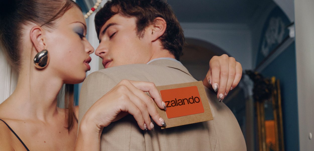 Fashion models share an intimate moment. One holds a brown card emblazoned with a red Zalando logo. Both sport striking accessories that complement their elegant outfits.