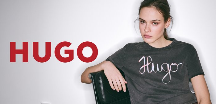 Graues Baumwoll-T-Shirt mit dem Schriftlogo "Hugo" in Pink, mit Rundhalsausschnitt und lockerer Passform, getragen im Sitzen vor einem schlichten Hintergrund.
