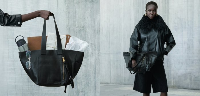 Femme en manteau en cuir noir avec col en fourrure tenant un grand sac cabas noir contenant une bouteille d'eau, des lunettes, un carnet et un mouchoir, appuyée contre un mur en béton.