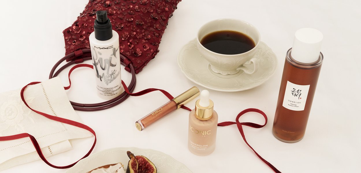 Taza de cerámica blanca con café, rodeada de productos para el cuidado de la piel: un frasco de spray transparente, dos sueros y un brillo labial sobre un mantel texturizado.