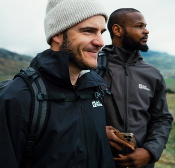 Zwei Männer im Freien, die Jacken tragen; einer in einer schwarzen wasserdichten Jacke mit Reißverschluss und Logo, der andere in einer braunen Jacke, beide mit Rucksäcken.