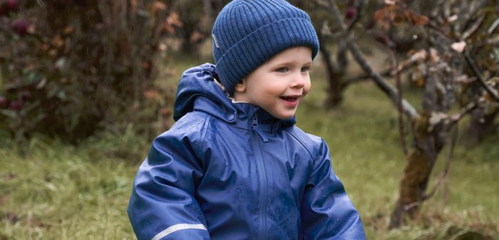 Kind in einem blauen wasserfesten Anzug mit reflektierenden Streifen, gestreiften Handschuhen und einer blauen Strickmütze, das auf einem hölzernen Gleichgewichtsrad auf Gras fährt.