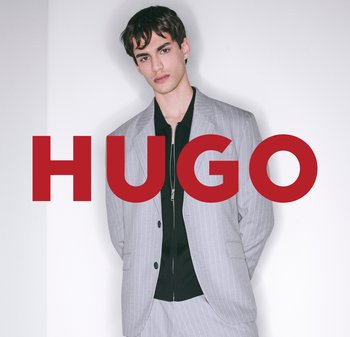 Veste de costume grise à rayures sur un chemisier noir, avec une fermeture éclair. Le modèle se tient devant un fond blanc, avec "HUGO" en rouge.