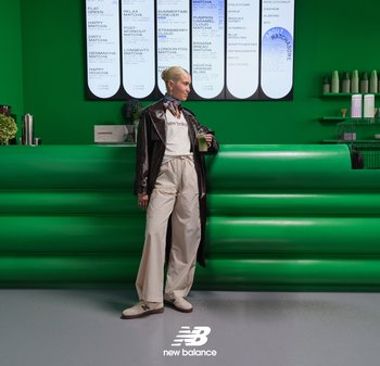 Mujer con una bebida verde se apoya en un mostrador verde brillante bajo un menú con opciones de bebida de matcha en una cafetería moderna.