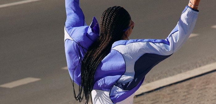 Personne aux longues tresses portant une veste bleue et blanche lève les deux bras en se tenant sur une rue ou un trottoir pavé.