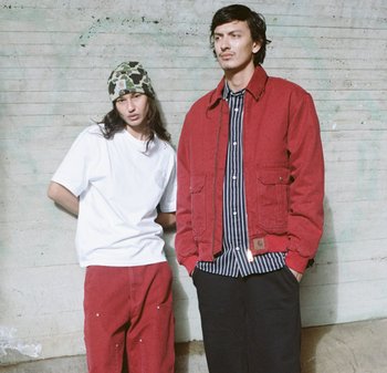 Dos modelos están de pie contra una pared texturizada. Uno lleva una camiseta blanca con pantalones rojos, el otro una chaqueta roja sobre una camiseta a rayas y pantalones oscuros.