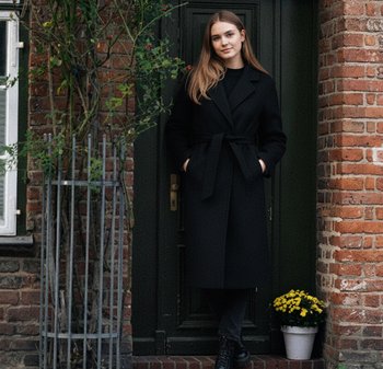 Palton din lână neagră cu centură, purtat peste un top și pantaloni negri. Stând pe trepte din cărămidă lângă o floare galbenă în ghiveci și o ușă verde.