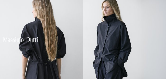 Vrouw met lang blond haar die een loszittende zwarte jas met knopen draagt, met handen in de zakken, tegen een effen lichte achtergrond.