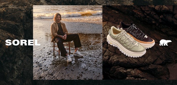 Man sitting on chair on wet sand wearing beige jacket and light sneakers, with two pairs of hiking shoes on dark rocks beside him.