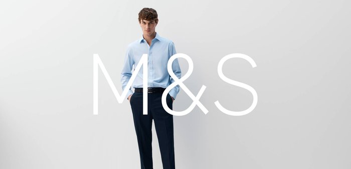 Young man wearing light blue button-up shirt and black trousers stands against plain gray background, M&S logo centered over him.
