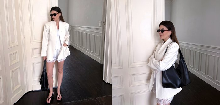 Femme portant un blazer blanc et un short avec des lunettes de soleil, portant un grand sac noir, debout dans un couloir avec des sols en bois foncé.