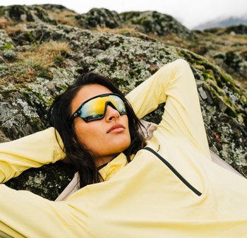 Schwarze Sonnenbrille mit reflektierenden gelben Gläsern, getragen von einer Person in einem hellgelben Langarmoberteil, die sich gegen felsiges Gelände lehnt.