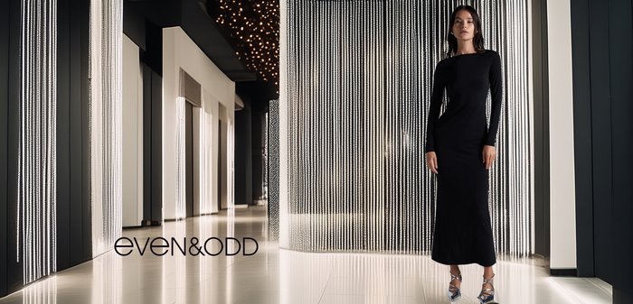 Zwarte jurk met lange mouwen en een strakke pasvorm, gestyled met elegante zilveren hakken. De achtergrond heeft fonkelende gordijnlichten en een modern interieurdesign.