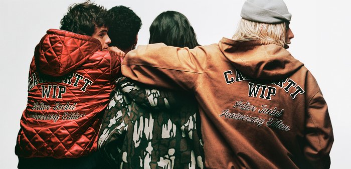 Chaqueta acolchada roja con la inscripción "Carhartt WIP Active Jacket Anniversary Edition" en la espalda, combinada con chaquetas con capucha en tonos marrones y estampado de camuflaje.