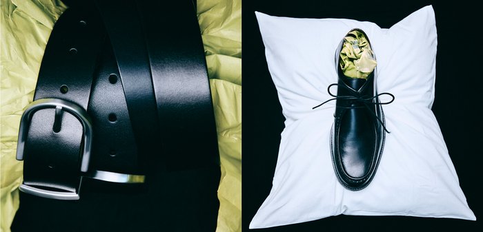 Schwarzer Ledergürtel mit silberner Schnalle und Löchern auf gelbem Hintergrund, daneben ein schwarzer Lederschuh auf einem weißen Kissen, gefüllt mit grünem Seidenpapier.