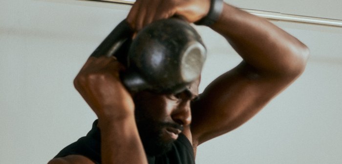 Moški dviguje kettlebell nad glavo, osredotočen na trening moči v minimalnem notranjem prostoru.