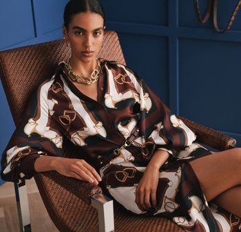 Vrouw met donker haar in een bruin-wit-blauw patroon jurk zit op een gevlochten stoel tegen een blauwe wand met panelen, en draagt een gouden ketting.