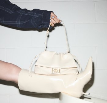 Jasnobeżowa torebka o miękkim, marszczonym designie z ozdobnym zapięciem. Dopasowana do błyszczących wysokich kozaków do kolan o ostrym nosku w tym samym kolorze.