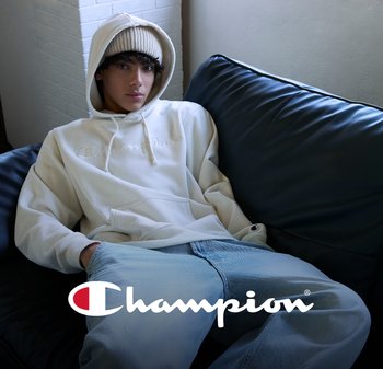 Giovane che indossa un hoodie bianco Champion e un berretto beige, seduto su un divano scuro, con il logo Champion sovrapposto al centro in basso.
