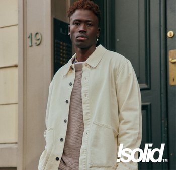 Beige Baumwolljacke mit Druckknöpfen, zwei Fronttaschen, über einem strukturierten leichten Pullover getragen. Das Modell posiert vor einer dunkelgrünen Tür.