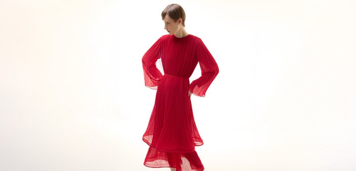 Femme portant une robe longue rouge plissée avec des manches longues transparentes et des talons ouverts noirs, debout les mains sur les hanches sur un fond blanc.