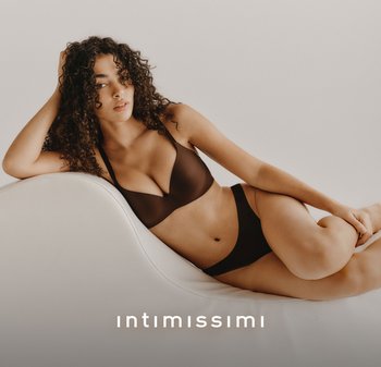 Donna con capelli ricci reclinata su una sedia curva bianca, indossa un reggiseno nero e biancheria intima davanti a uno sfondo beige uniforme.