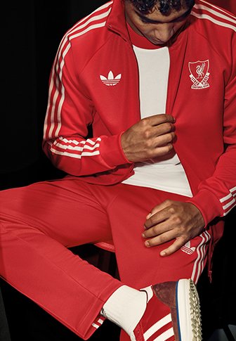 Personne portant une veste de survêtement rouge Adidas Liverpool, ajustant la veste, assise avec une jambe croisée sur l'autre.