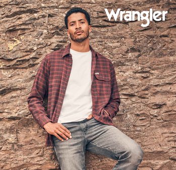 Rød og svart rutete skjorte over en hvit t-skjorte, kombinert med grå jeans. Modell poserer foran en teksturert steinvegg. Wrangler-logo synlig.