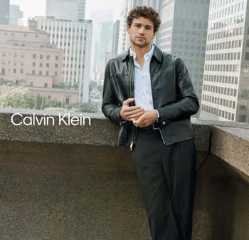 Joven con chaqueta de cuero negra y camisa blanca apoyado en una barandilla de concreto, con rascacielos de la ciudad al fondo.