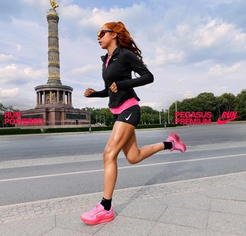 Abbigliamento da corsa Nike che include una giacca nera con zip, una canotta rosa e pantaloni corti neri. Il corridore indossa scarpe da ginnastica rosa e occhiali da sole. Monumento a colonne sullo sfondo.