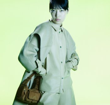 Beżowa skórzana koszula i spodnie, z nałożonym na to oversize'owym, jasnoszarym wełnianym płaszczem. Brązowa torba na ramię z kremowymi akcentami, ustawiona na limonkowym tle.