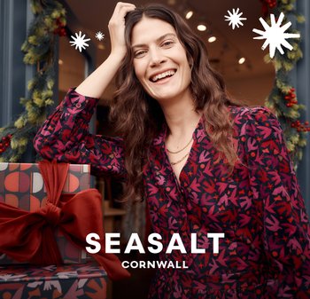 Šaty s květinovým vzorem v tmavě červených a fialových tónech, doplněné vrstvenými zlatými náhrdelníky, na pozadí zabalených dárků a slavnostní výzdoby.