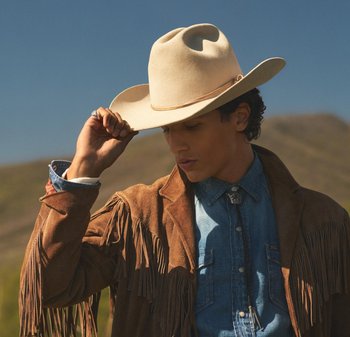 Giovane all'aperto che solleva un cappello da cowboy beige, indossando una giacca in suede marrone con frange e una camicia di denim con cravatta a bolo, contro un cielo blu chiaro.