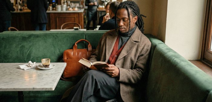 Homme aux cheveux tressés portant un manteau marron, lisant un livre assis sur un banc en velours vert près d'une fenêtre dans un café, avec une tasse de café et un sac à proximité.