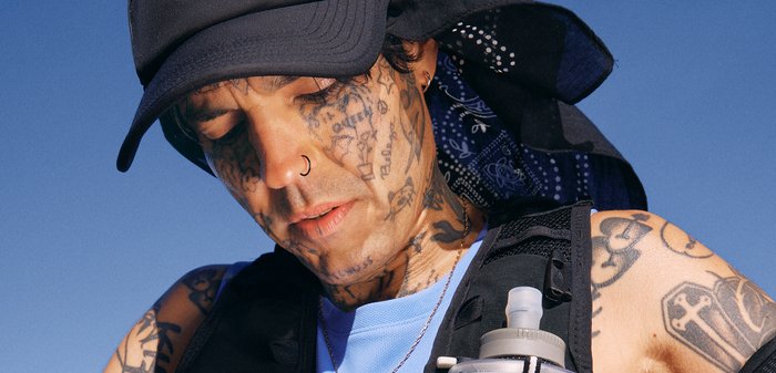 Personne avec de nombreux tatouages sur le visage et les bras porte une casquette et un bandana noirs, un anneau nasal, et un gilet noir avec une bouteille d'eau, à l'extérieur sous le soleil.