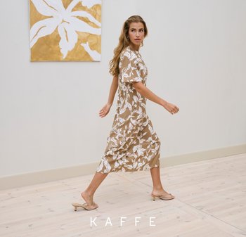 Woman walking indoors wearing a beige floral midi dress and beige heeled sandals on light wooden floor with gold and white artwork.