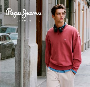 Young man wearing a red sweatshirt and headphones around his neck walks on a city sidewalk near a glass storefront.