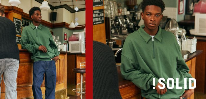 Grüner Pullover mit Reißverschlusskragen, lockere Passform, getragen über blauer Jeans. Bar mit Holzakzenten und Glasgeschirr im Hintergrund.